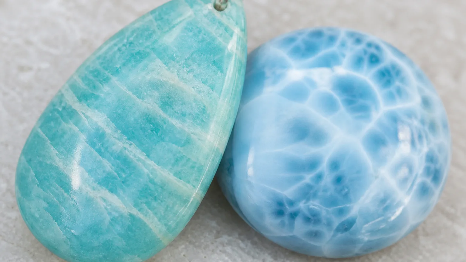 A macro close-up of blue-green Amazonite (pendant) and blue Larimar (cabochon pendant) crystals arranged together in one frame, both stones touching, fine surface textures visible, soft even studio light