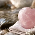 A soft pink rose quartz heart-shaped stone resting on a folded cloth near gentle flowing water, soft diffused light, tender calm mood