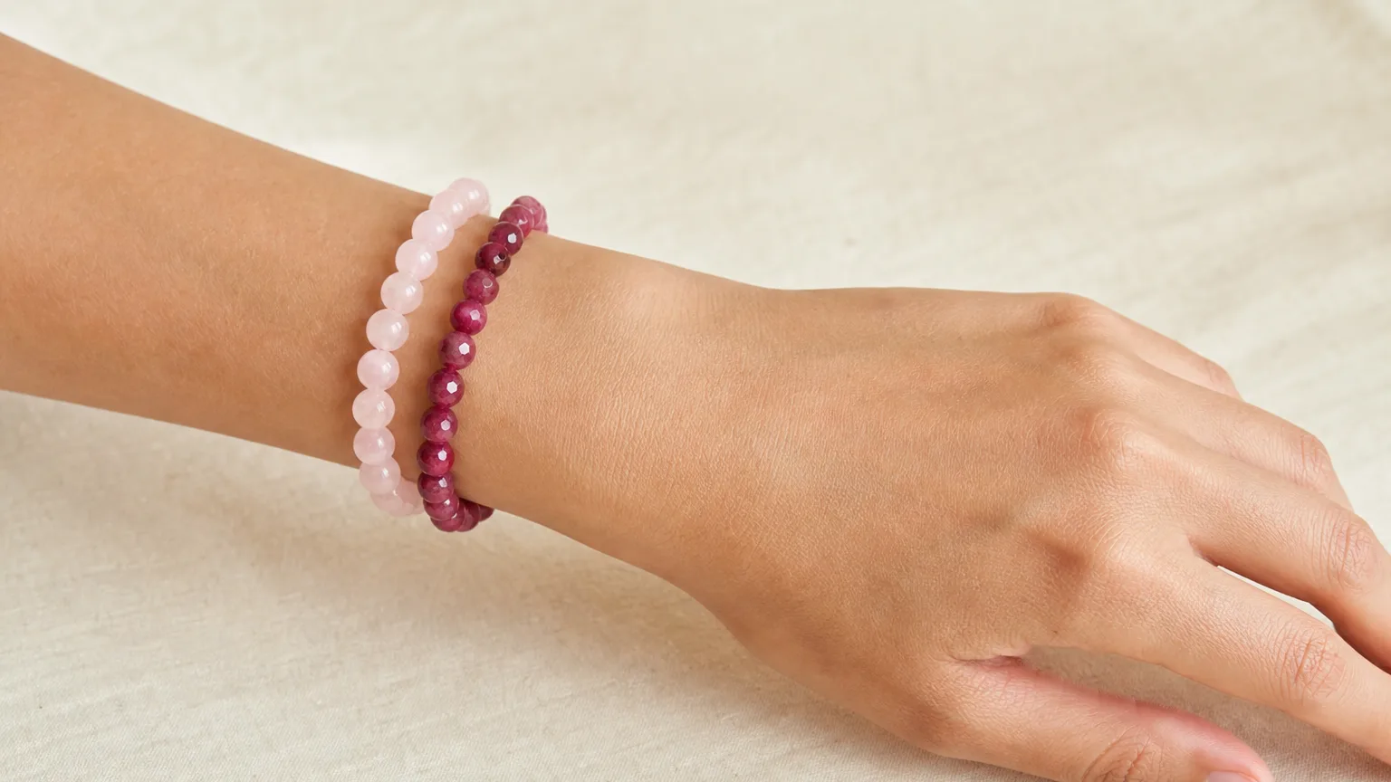 Rose Quartz and Ruby crystal bracelets worn together stacked on one wrist, real hand, natural daylight, minimalist wellness lifestyle scene