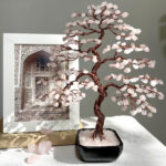 May include: A pink rose quartz wire wrapped tree with copper wire branches. The tree is in a black pot with a base of pink rose quartz crystals.