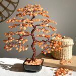 May include: A copper wire tree sculpture with carnelian stones. The tree is in a black pot and is surrounded by small carnelian stones. The tree is on a white surface.