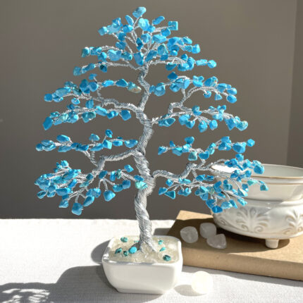 May include: A decorative bonsai tree sculpture with a silver wire trunk and branches. The branches are adorned with small, bright blue gemstones, resembling leaves. The tree sits in a square white ceramic pot filled with clear stones.