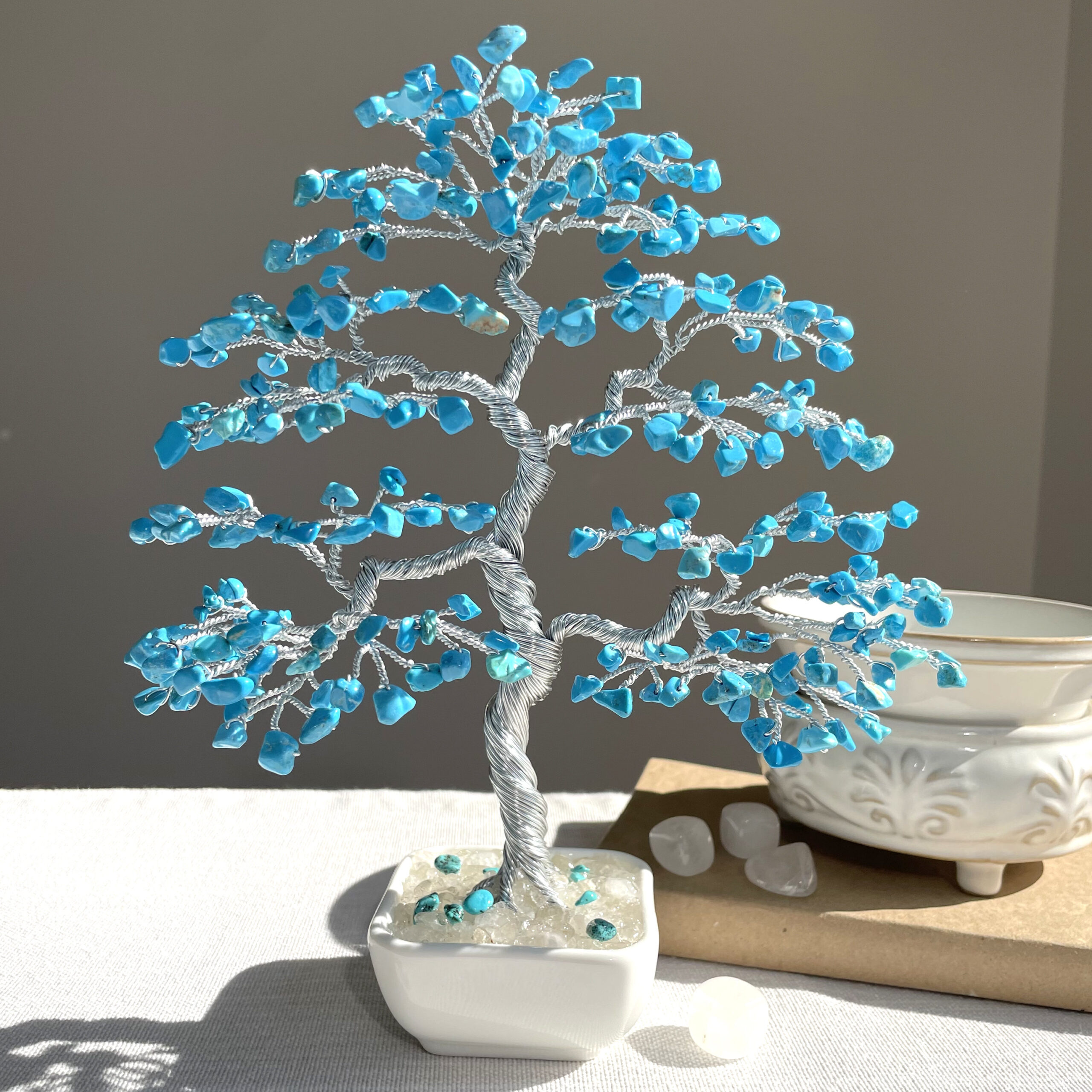 Natural Turquoise Bonsai Tree - Image 1 May include: A decorative bonsai tree sculpture with a silver wire trunk and branches. The branches are adorned with small, bright blue gemstones, resembling leaves. The tree sits in a square white ceramic pot filled with clear stones.