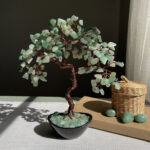 May include: A decorative tree sculpture with a copper wire trunk and branches, adorned with green gemstone leaves. The tree sits in a black bowl filled with matching stones. A small woven basket and three green stones are next to the tree.