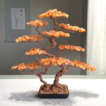 May include: A decorative bonsai tree sculpture. The tree features a twisted copper wire trunk and branches, adorned with orange-colored gemstone leaves. The tree sits in a black rectangular pot filled with small stones. The sculpture is a unique home decor item.