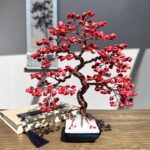 May include: A red coral wire wrapped bonsai tree in a black pot. The tree is made of copper wire and red coral beads. The tree is sitting on a wooden table.