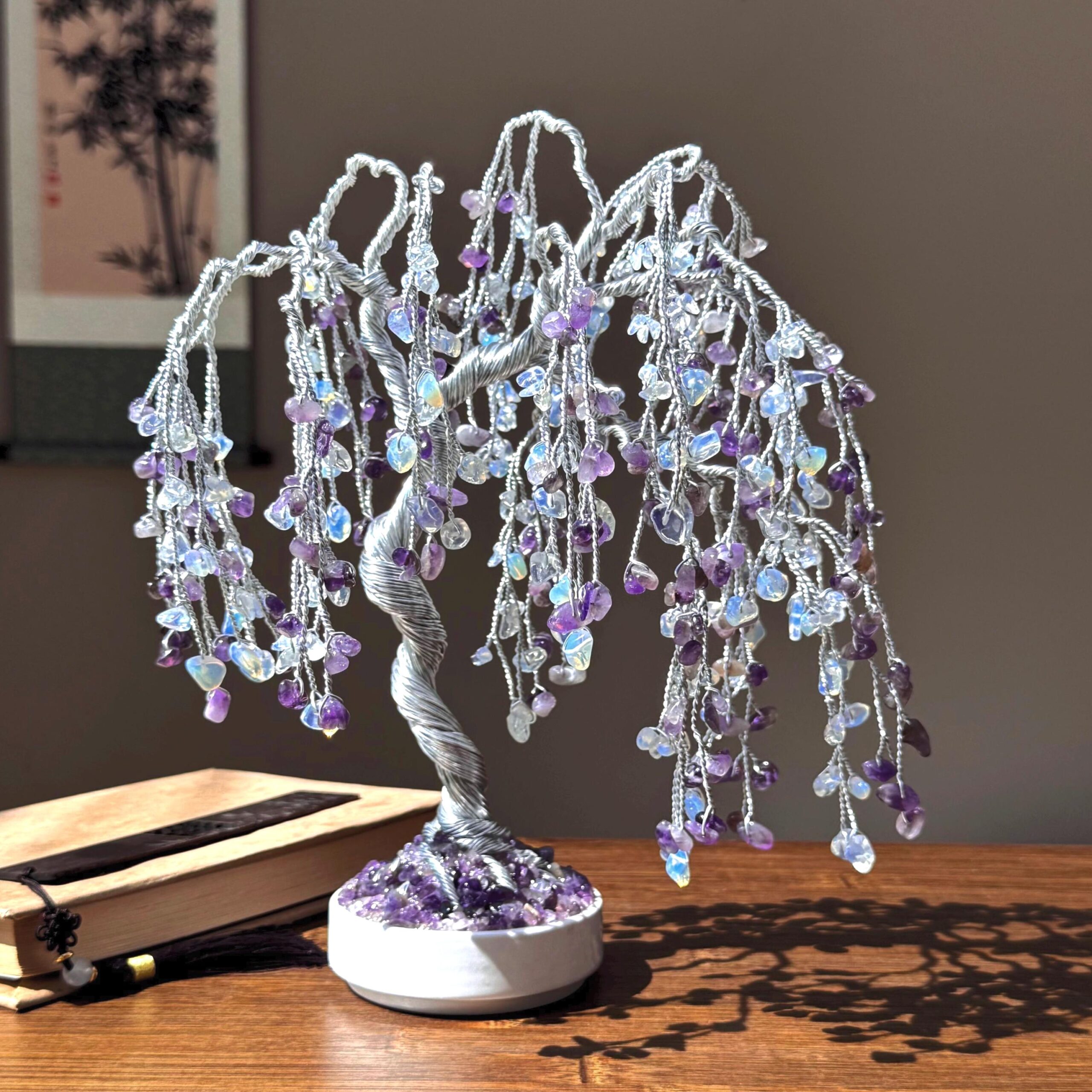 Natural Amethyst + Amethyst And Opal Willow Tree - Image 1 May include: A silver wire tree sculpture with amethyst and clear crystal stones hanging from the branches. The tree is in a white ceramic pot with a base of amethyst stones.