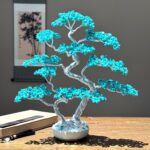 May include: A decorative bonsai tree sculpture with a silver wire trunk and branches. The leaves are made of small, turquoise-colored stones. The tree sits in a white ceramic pot on a wooden surface. A scroll painting is in the background.