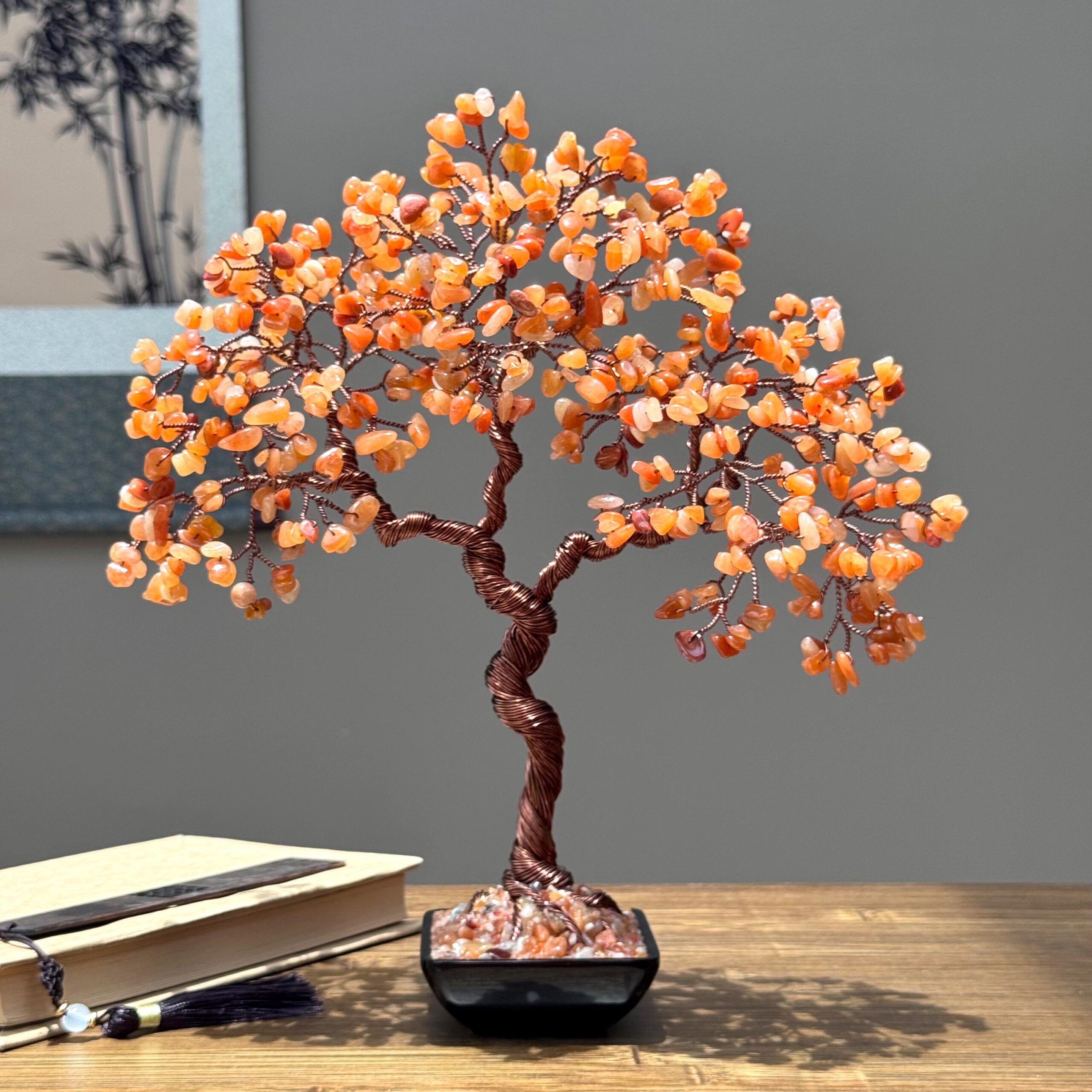 Natural Red Aventurine Crystal Tree - Image 1 May include: A decorative gemstone tree with a twisted copper wire trunk and branches. The tree's leaves are made of small, orange-colored gemstones. It sits in a square, black pot on a wooden surface. A book with a tassel is in the foreground.