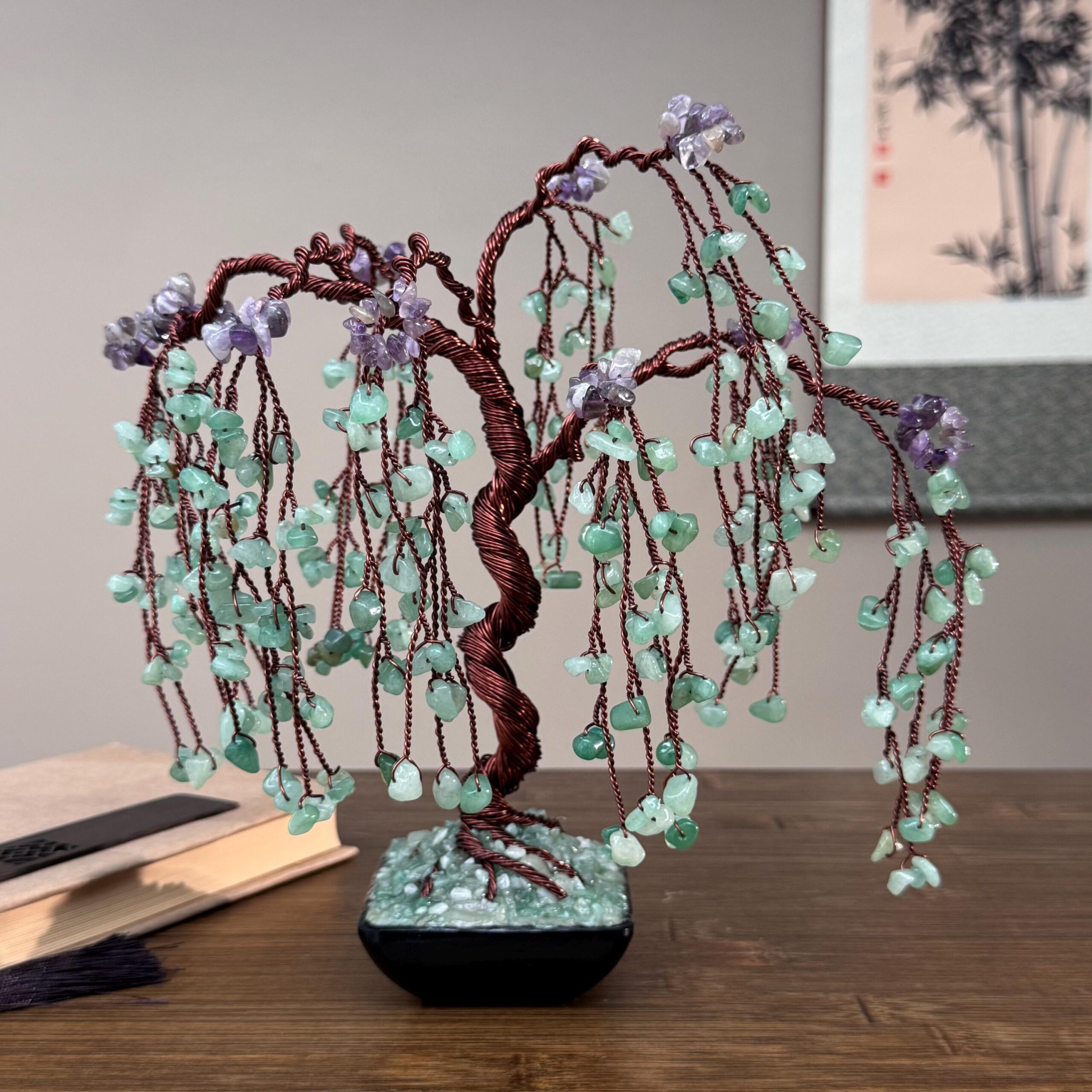 Natural Aventurine + Aventurine And Aquamarine Bonsai Tree (Copper Wire) - Image 1 May include: A decorative wire tree sculpture with a twisted brown trunk and branches. The branches are adorned with green and purple gemstone leaves and flowers. The tree sits in a black square pot on a wooden surface.
