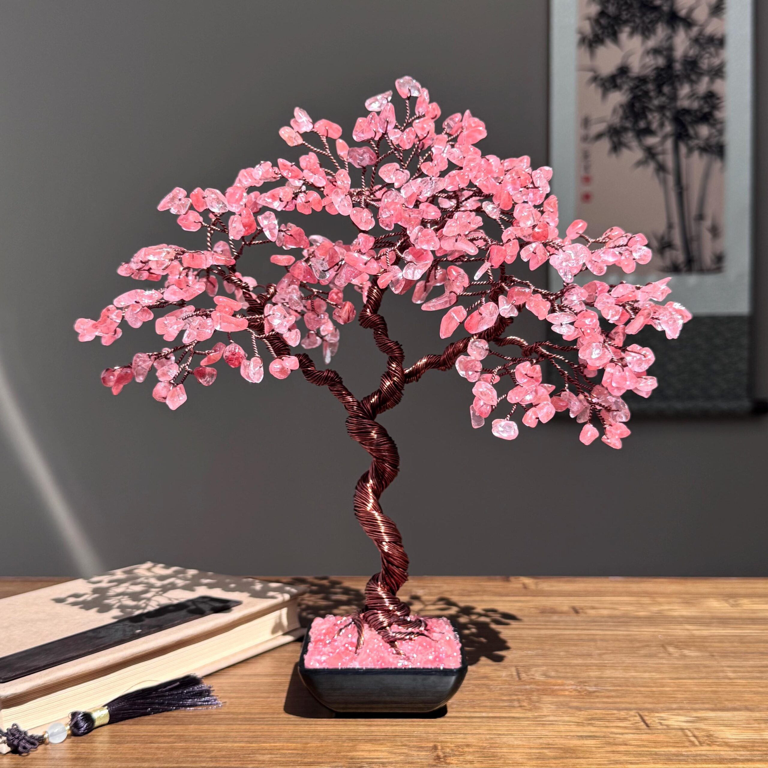 Handmade Cherry Quartz Tree of Life - Image 1 May include: A decorative bonsai tree sculpture with a twisted copper wire trunk and branches. The tree's leaves are made of pink gemstones, and it sits in a black square pot. The tree is on a wooden surface.