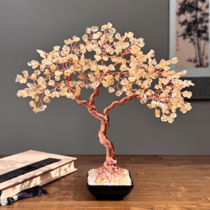 May include: A decorative tree sculpture with a copper wire trunk and branches, adorned with amber-colored crystal chips. The tree sits in a square black pot filled with crystals. The sculpture is on a wooden surface.