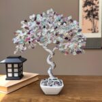 May include: A decorative tree sculpture with a twisted silver wire trunk and branches. The tree's leaves are made of colorful, polished gemstones in shades of purple, green, and clear. The tree sits in a square white ceramic pot.