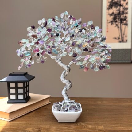 May include: A decorative tree sculpture with a twisted silver wire trunk and branches. The tree's leaves are made of colorful, polished gemstones in shades of purple, green, and clear. The tree sits in a square white ceramic pot.