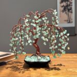 May include: A decorative tree sculpture featuring a twisted copper wire trunk and branches adorned with small, light green tumbled stone leaves. The tree is anchored in a black rectangular ceramic pot filled with matching green stone chips.
