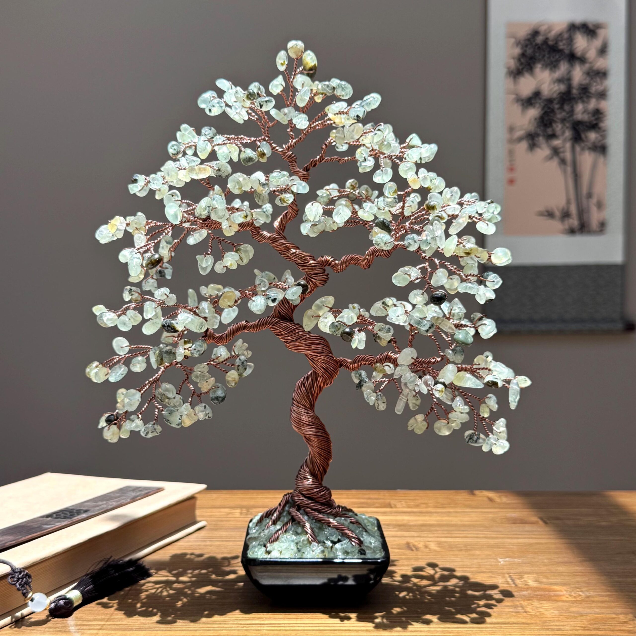 Natural Prehnite Bonsai Tree - Image 1 May include: A decorative wire tree sculpture featuring a twisted copper-colored trunk and branches adorned with numerous small, pale green translucent stone chips. The tree is anchored in a square black pot filled with matching green stone chips.