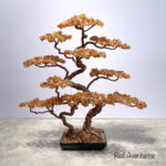 May include: A decorative bonsai tree sculpture made with copper wire and red aventurine gemstones. The tree has a twisted trunk, branches, and a square black base. The gemstones are a warm orange color. Text at the bottom reads "Red Aventurine".