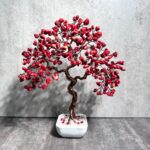 May include: A red coral wire wrapped bonsai tree in a white ceramic pot. The tree is made of copper wire and red coral beads. The pot is sitting on a gray surface.