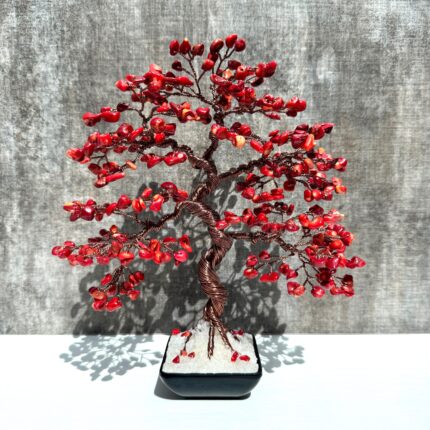 May include: A red coral wire wrapped bonsai tree in a black pot. The tree is made of copper wire and red coral beads. The tree is sitting on a white surface.
