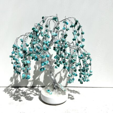 May include: A handcrafted wire tree sculpture with a silver-colored trunk and branches. The branches are adorned with turquoise-colored gemstones, creating a weeping willow effect. The tree sits on a white base.