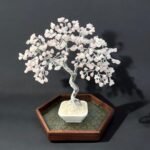 May include: A decorative wire tree sculpture with a twisted trunk and branches adorned with pink gemstones. The tree sits in a white square pot on a wooden tray with a hexagonal shape. The overall design is elegant and artistic.