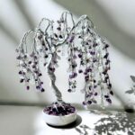 May include: A silver wire tree sculpture with amethyst and moonstone crystals. The tree has a weeping willow shape and is displayed on a white base.
