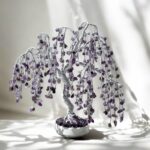 May include: A silver wire tree sculpture with amethyst crystals hanging from the branches. The tree is in a white pot and is on a white surface.