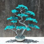 May include: A handcrafted bonsai tree sculpture with a silver wire frame and vibrant turquoise stone leaves. The tree is set in a white ceramic pot, creating a striking contrast against the gray background. The sculpture casts a shadow.