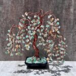 May include: A copper wire bonsai tree sculpture with a twisted trunk and cascading branches. The branches are adorned with small, light green and yellow gemstone chips. The tree sits in a black square pot filled with green stones, casting a shadow.