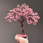 May include: A handcrafted wire tree sculpture with a twisted copper trunk and branches. The tree's leaves are made of pink gemstones, and it sits on a black base. The sculpture is held up by a hand.