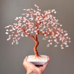 May include: A handcrafted copper wire tree sculpture with clear crystal gemstone leaves. The tree is in a white square pot filled with small, clear stones. The tree is held in a hand against a neutral background.