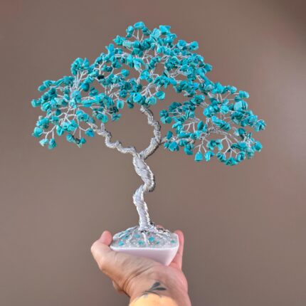 May include: A handcrafted wire tree sculpture with turquoise-colored stone leaves. The tree has a silver-colored twisted wire trunk and branches, and is set in a white square base. The sculpture is held in a hand.