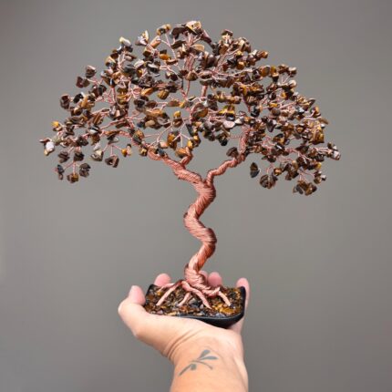 May include: A decorative wire tree sculpture featuring a twisted copper-colored trunk and branches adorned with numerous small, polished brown and gold tiger's eye gemstone chips. The base is a black rectangular container filled with stone chips.