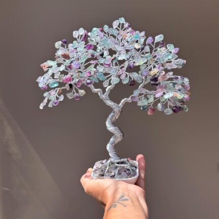 May include: A handcrafted wire tree sculpture with a twisted silver trunk and a canopy of colorful, polished gemstones. The gemstones are in shades of purple, green, and pink. The tree is set in a white square base.