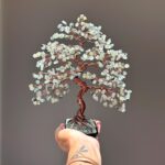May include: A decorative wire tree sculpture featuring a twisted copper-colored metal trunk and branches adorned with numerous small, pale green tumbled gemstone chips. The base is a square black container filled with matching green stone fragments.