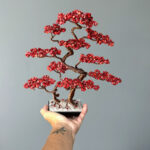 May include: A red wire-wrapped bonsai tree with a copper wire frame and red gemstone leaves. The tree is displayed on a black base with white stones.