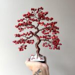May include: A red wire-wrapped bonsai tree with red gemstone leaves. The tree is in a small black square pot with white stones.