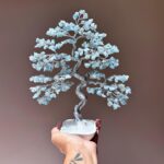 May include: A wire-wrapped crystal tree with aquamarine stones, a white base, and a twisted trunk. The tree is held in a hand with a dragonfly tattoo.