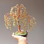 May include: A copper wire sculpture of a weeping willow tree with green gemstone leaves. The tree is in a black pot with green moss.